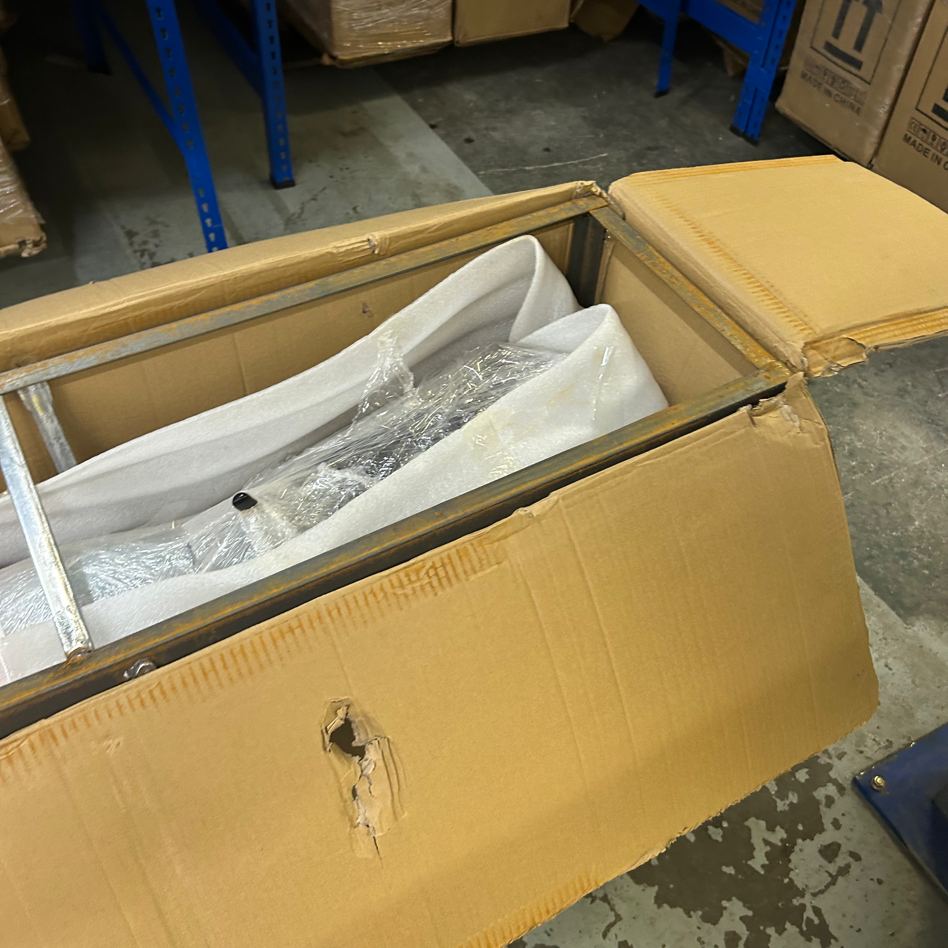 Open cardboard box with a wrapped scooter inside, on a concrete floor with boxes in the background.