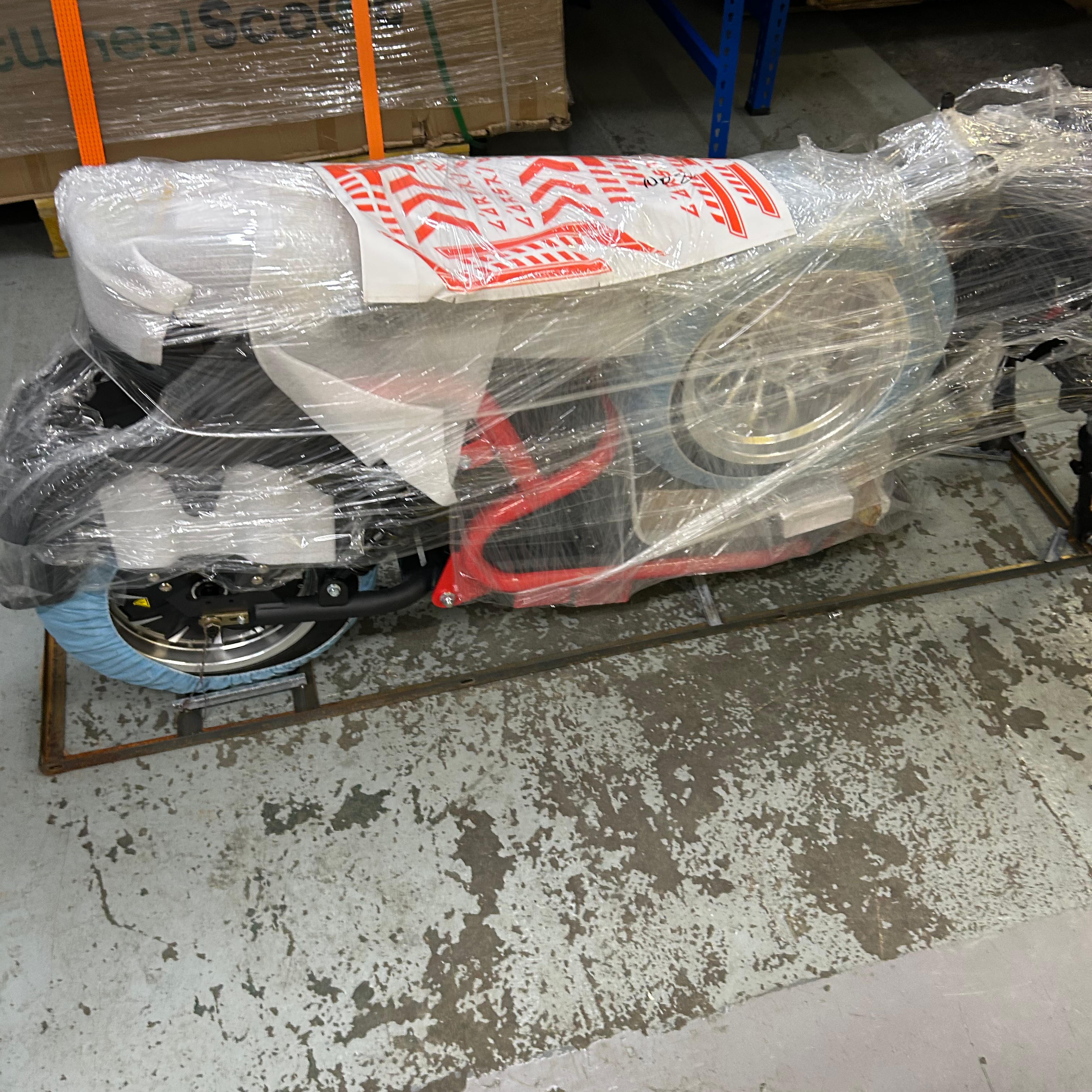 Wheeled scooter wrapped in plastic with red and white label in a warehouse setting